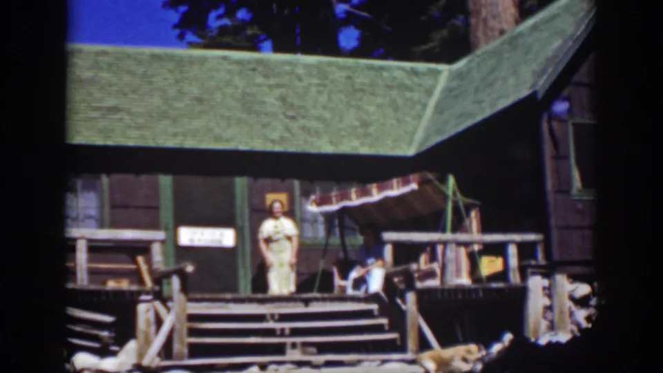 Lake Tahoe 1939 frame with a woman in a light dress on a wooden cabin porch, seated person nearby, and pine trees behind the roof.