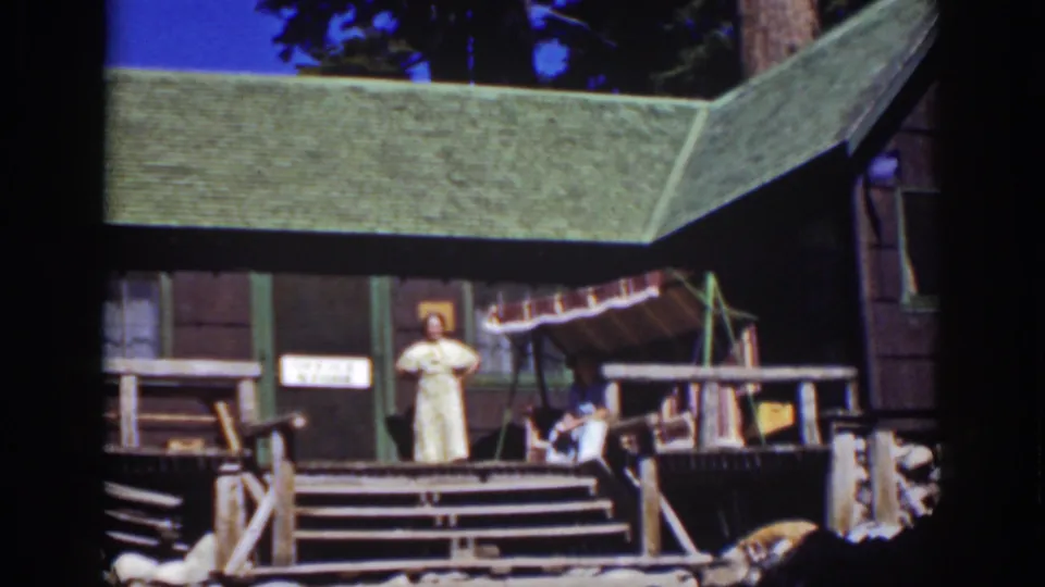 Lake Tahoe 1939 home-movie frame of a woman on a rustic cabin porch, with wood steps, railing, awning, and a seated figure nearby.