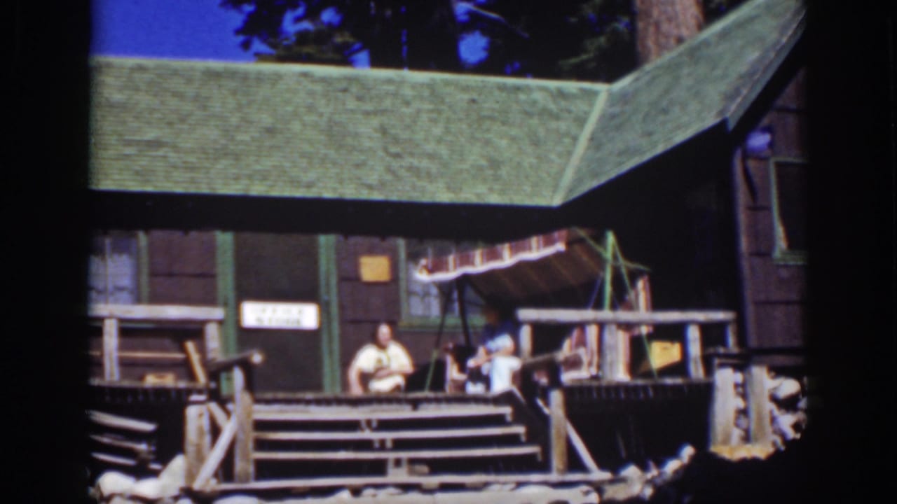 Wide view of a rustic cabin porch with people, a striped canopy, steps, and a partial office-store sign.