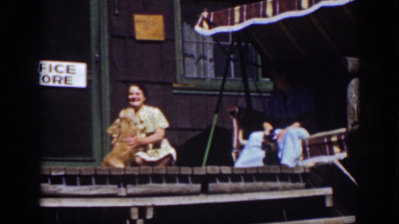 Woman seated on a wooden porch with a golden dog beside her and another adult in a striped swing.