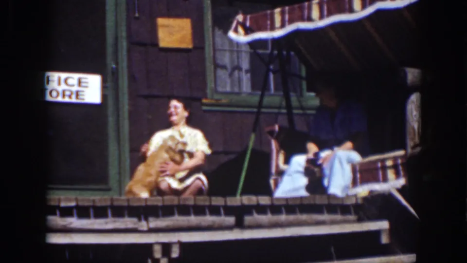 1939 Lake Tahoe porch moment with a woman handling a golden dog, a second adult in a swing, and the partial “OFFICE STORE” sign behind them.