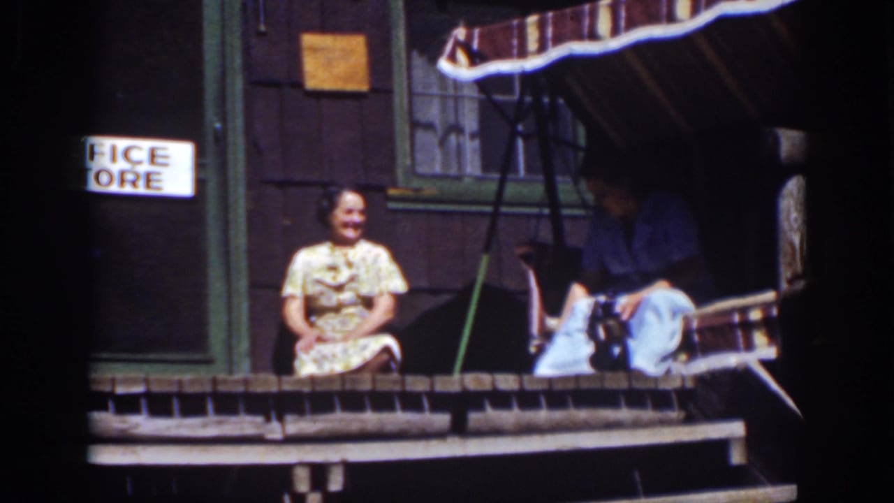 Two adults sit on a wooden porch outside a dark cabin with a striped awning.