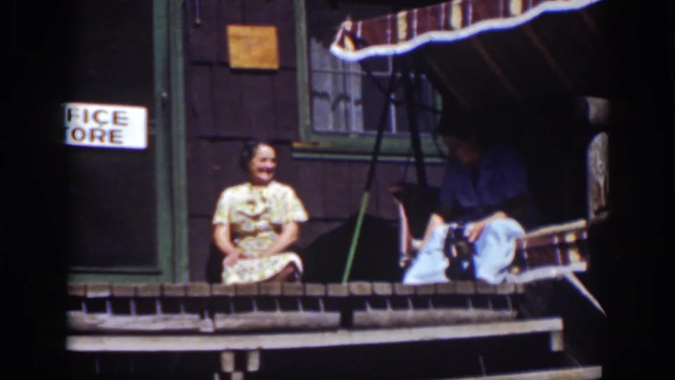 1939 Lake Tahoe home-movie frame showing a smiling woman and seated man on a wooden porch beside a cabin entrance.