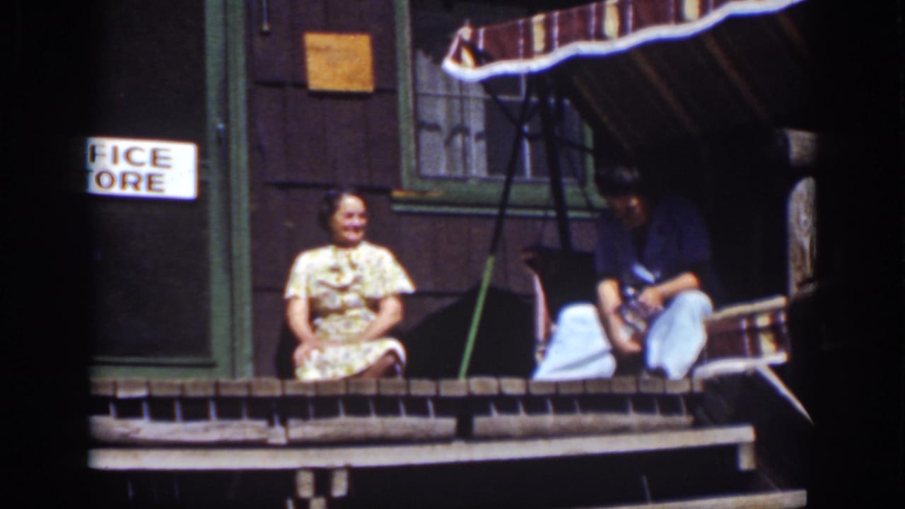 A woman smiles on a porch while a man sits beside her near a cabin doorway.