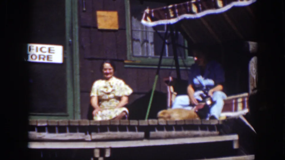 1939 Lake Tahoe frame showing two adults on a wooden cabin porch, with a striped awning and partial office-store sign nearby.
