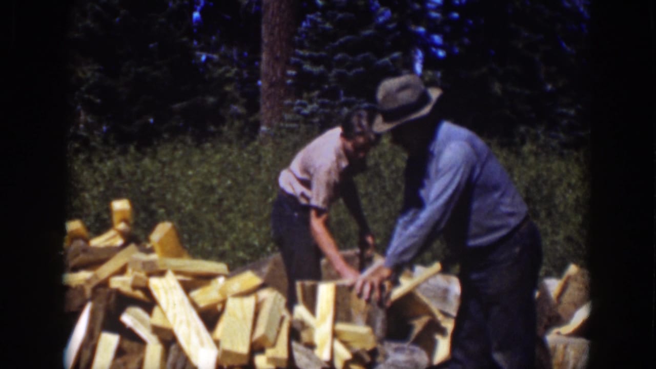 1939 Lake Tahoe frame with two people coordinating around an upright log and axes at a forest woodpile.