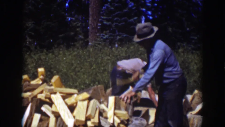 Final 1939 Lake Tahoe frame with the younger figure bending at the woodpile as the older man steadies a log in the wooded setting.