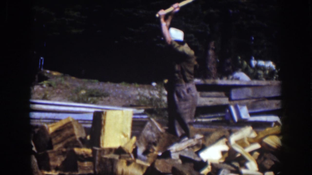 Man in a white hat raises an axe above a chopping block beside split wood.
