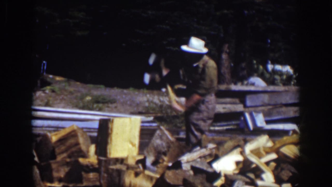 Man bends forward with an axe near a chopping block and woodpile.