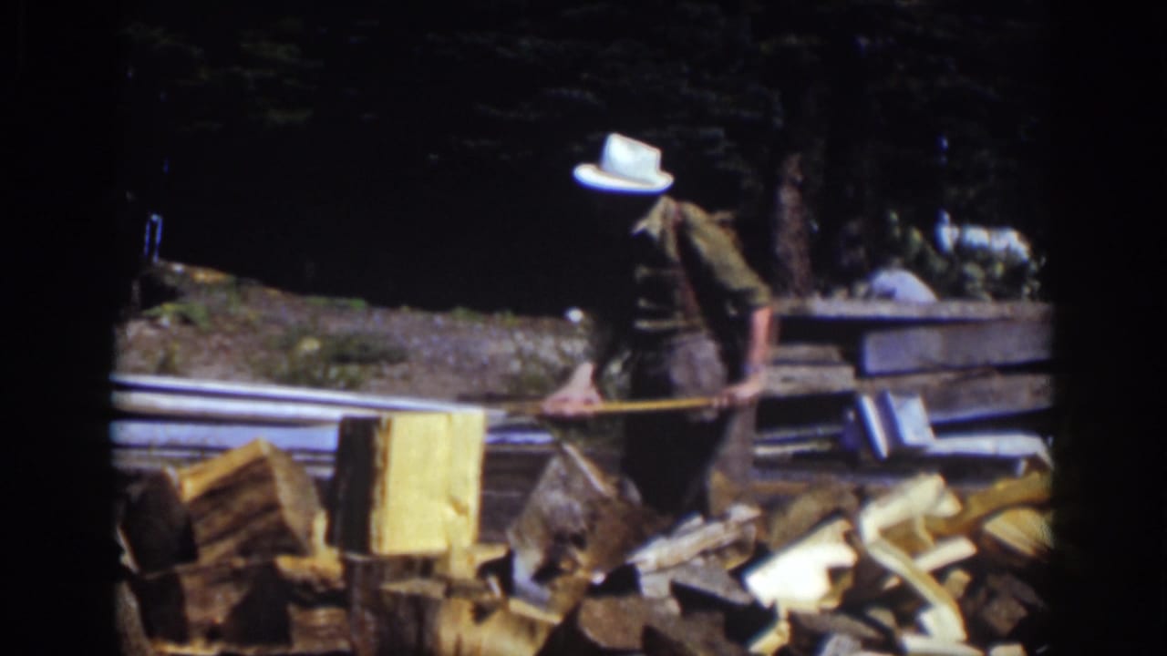 Man in a white hat bends over a chopping area beside a pile of split wood.
