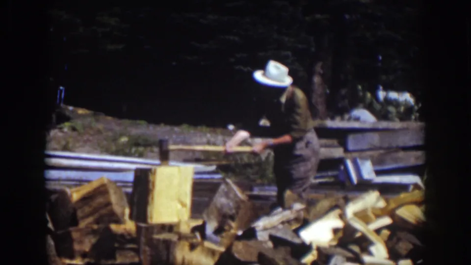 A man in a white hat stands outdoors at Lake Tahoe with an axe, surrounded by split wood and boards.