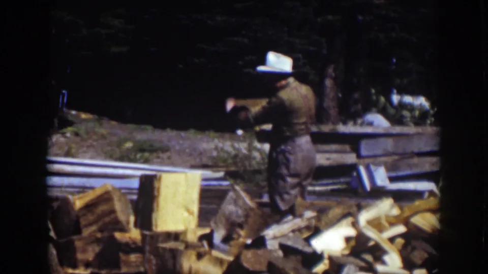 In the final frame, the man faces partly away with the axe raised near a pile of split wood and stacked lumber.