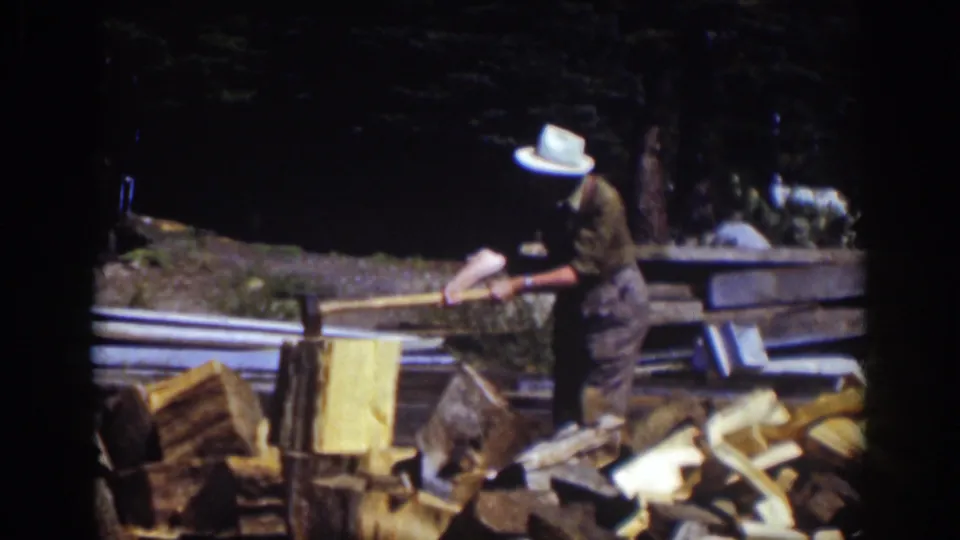 At Lake Tahoe in 1939, a man in a white hat swings an axe near stacked logs and rough lumber in a wooded outdoor area.