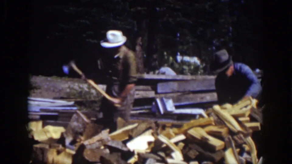 Two men continue firewood work at Lake Tahoe in 1939, with axes, logs, and rough boards visible in a sunlit wooded setting.
