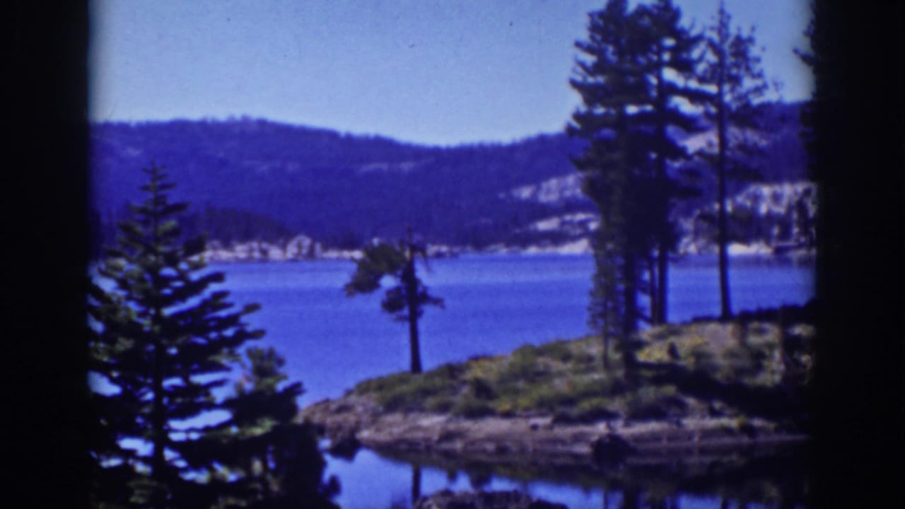 Pine trees and a small rocky shore beside a blue lake with wooded hills beyond.