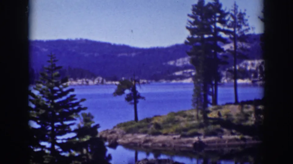 Soft 8mm view of Lake Tahoe with a pine-lined rocky shore, calm water, and wooded hills in the distance.