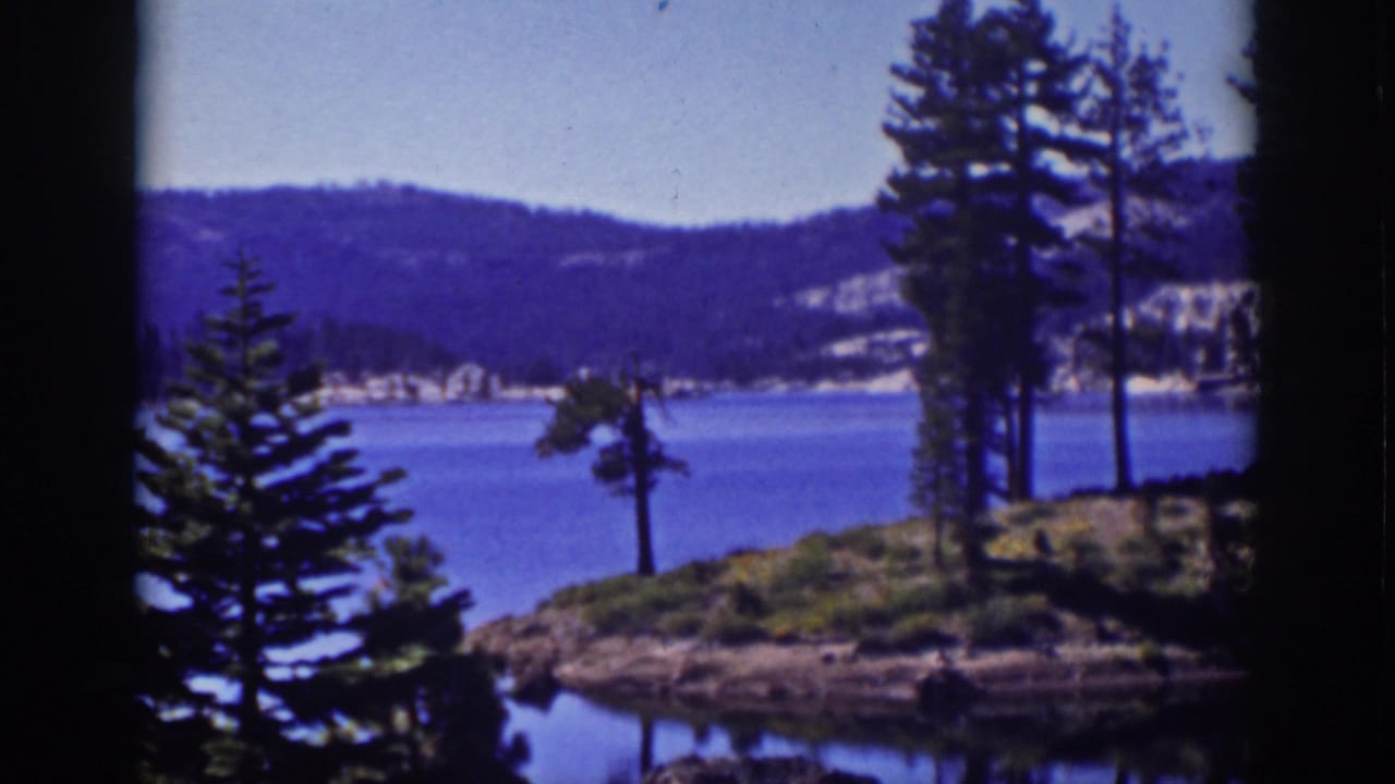 Blue lake with pine trees, rocky shore, distant hills, and dark 8mm frame borders.