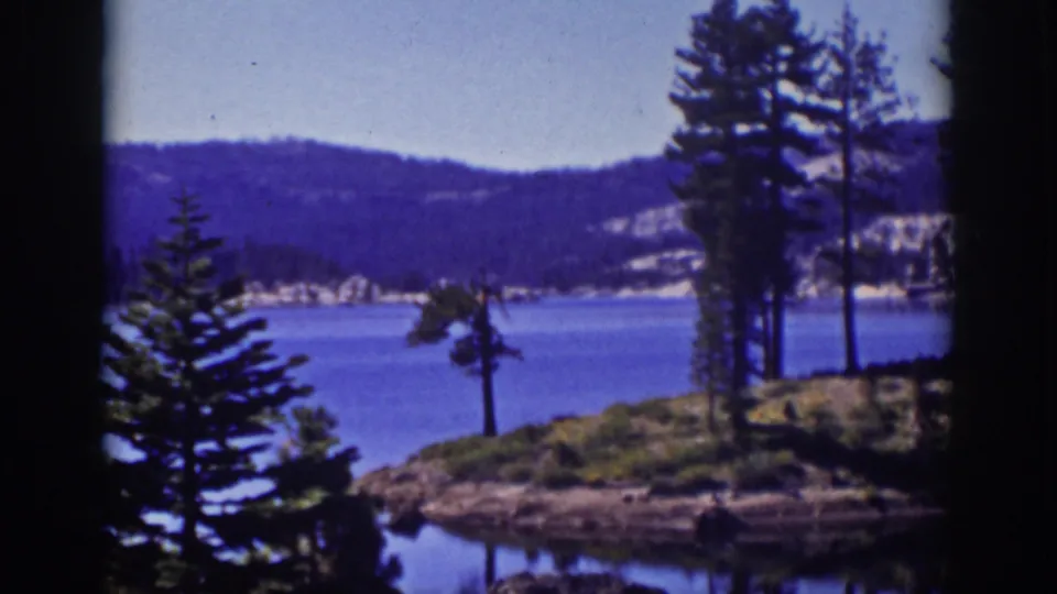 1939 Lake Tahoe scene with pines along a rocky point, blue water, distant wooded hills, pale sky, and visible 8mm frame edges.