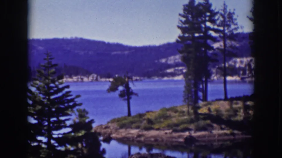 1939 Lake Tahoe 8mm frame showing calm water, a pine-lined rocky shore, wooded hills in the distance, and soft grainy color.
