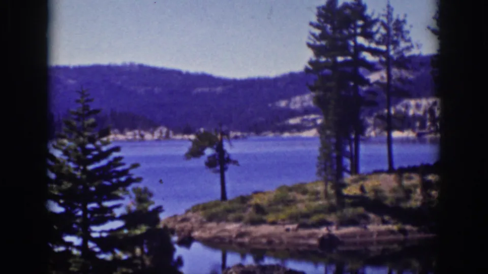 1939 Lake Tahoe home-movie frame with a pine-lined rocky shore in the foreground, blue lake water, distant hills, and rounded projection borders.