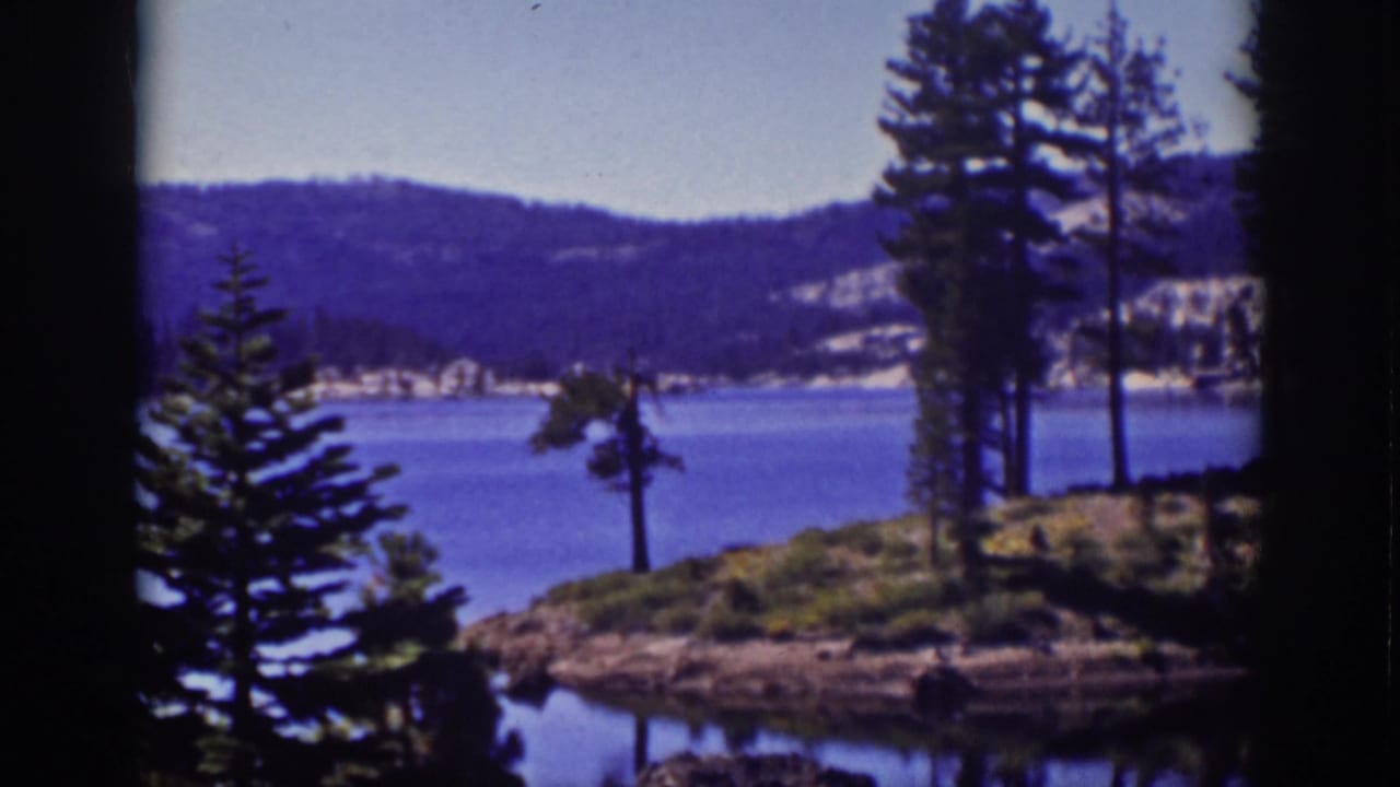 Lake shoreline with pine trees, blue water, distant hills, and dark film borders.