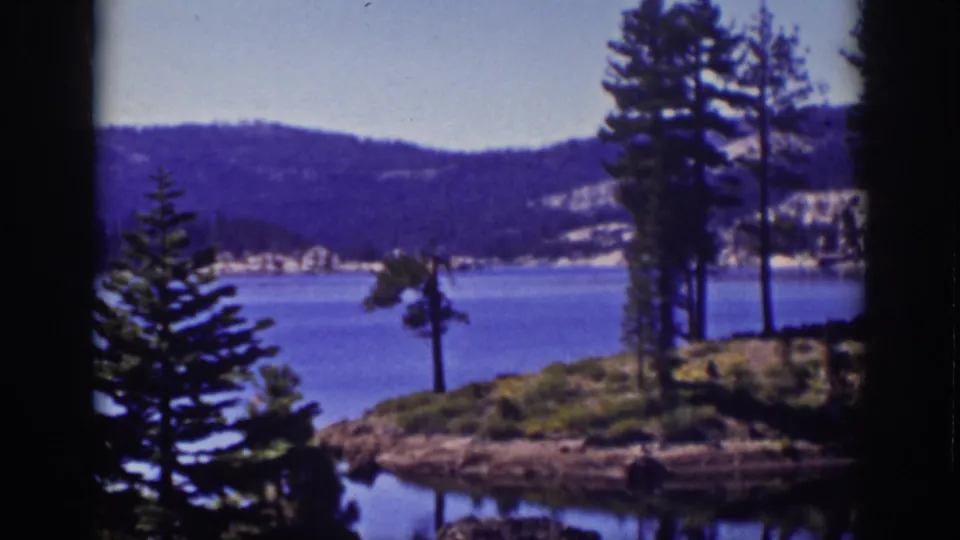 1939 Lake Tahoe home-movie frame showing pine trees on a rocky shore, blue water, distant forested hills, and dark 8mm frame edges.