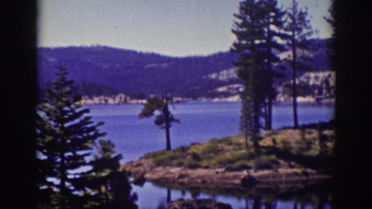 Blue lake framed by dark tree silhouettes, pine trees, rocky shoreline, and distant hills.