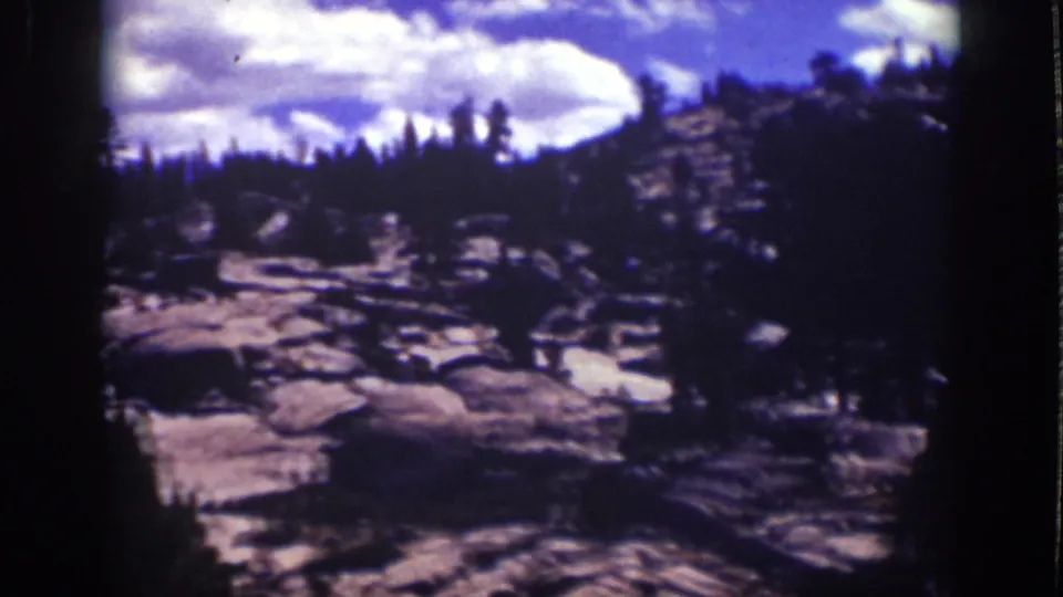 Archival Lake Tahoe frame showing the pan across granite-like rocks, scattered pines, shaded trees, and bright clouds.