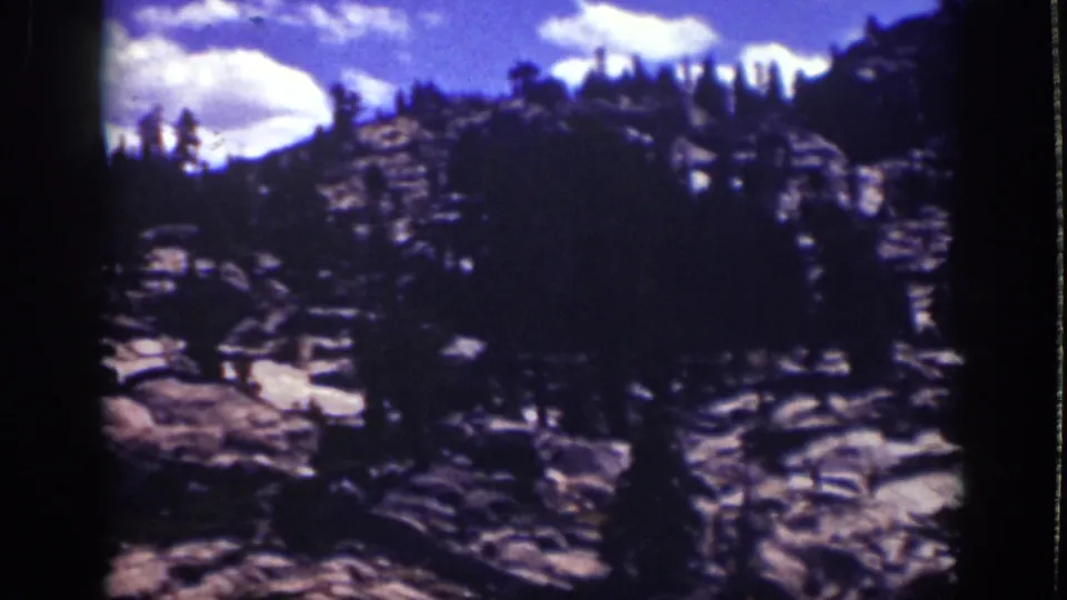 Late-frame view of Lake Tahoe mountain scenery, with dense dark pines partly obscuring rock slabs beneath a bright clouded sky.