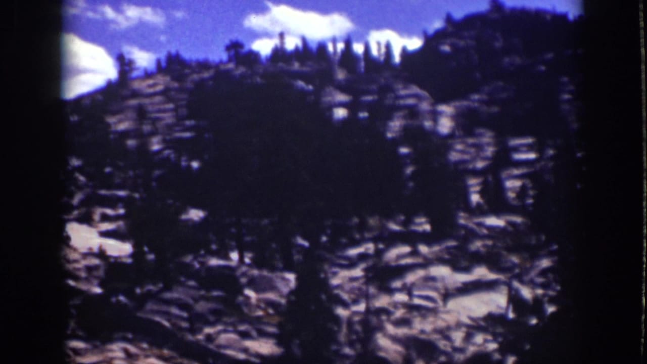 Rocky forested hillside with pine trees beneath blue sky and white clouds.