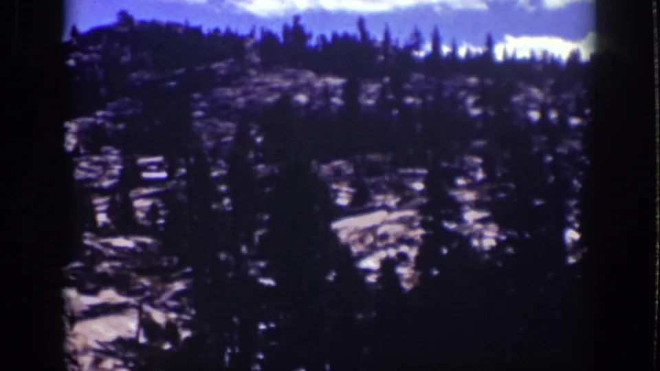 1939 Lake Tahoe frame showing a forested rocky ridge, dark pine outlines, pale stone surfaces, and aged blue-purple color.