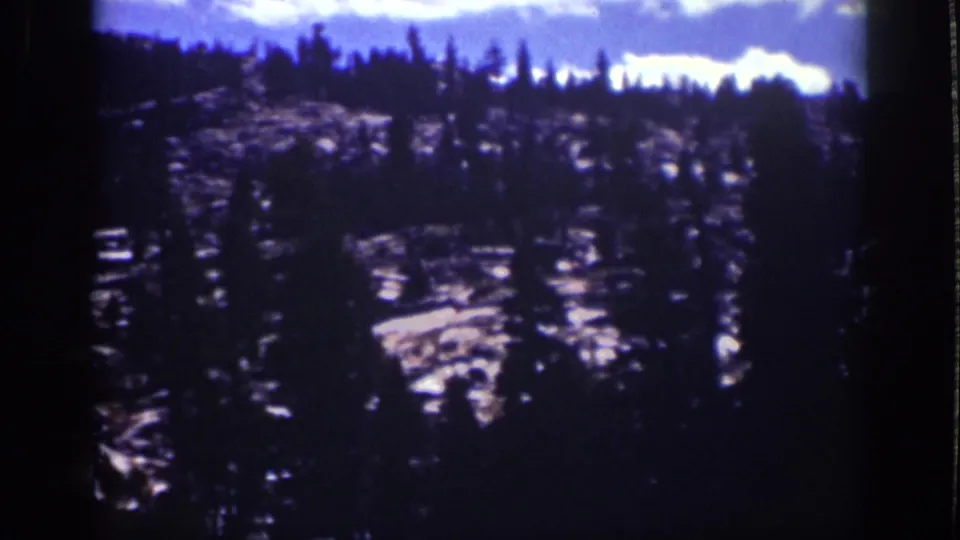 1939 Lake Tahoe 8mm frame showing dark pine trees across a rocky hillside under a blue sky with pale clouds.