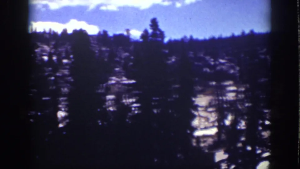 Vintage Lake Tahoe frame showing a pine-dotted rocky hillside, dark tree silhouettes, and a bright blue sky with cloud shapes.
