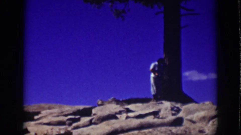 1939 Lake Tahoe frame showing an adult in light clothing beside a large pine trunk on rocks against a deep blue sky.