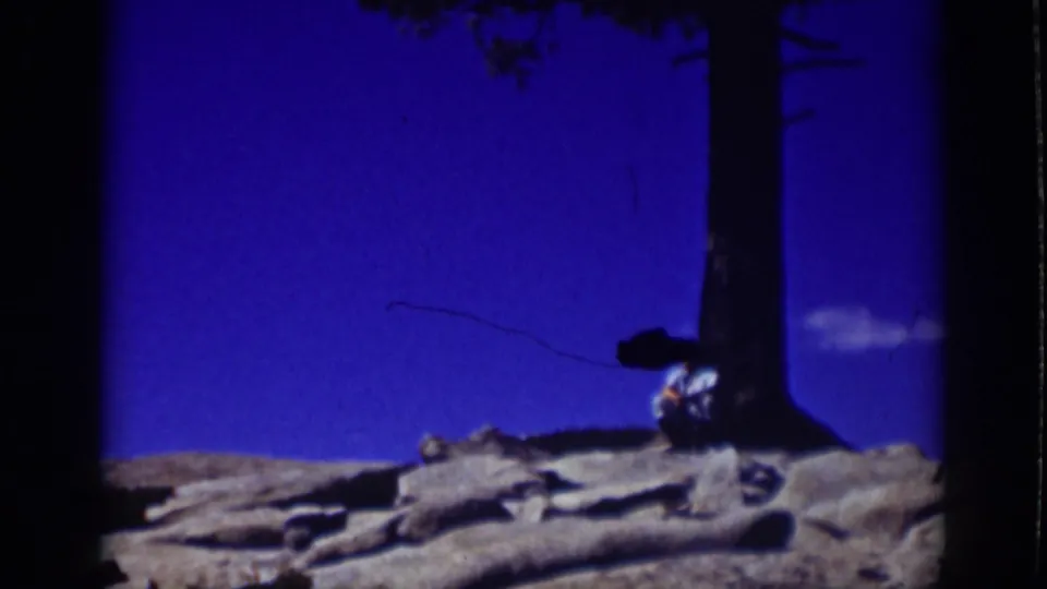 Blue-tinted 1939 Lake Tahoe home-movie frame showing the seated person near the tree with rocks and shoreline forms below.