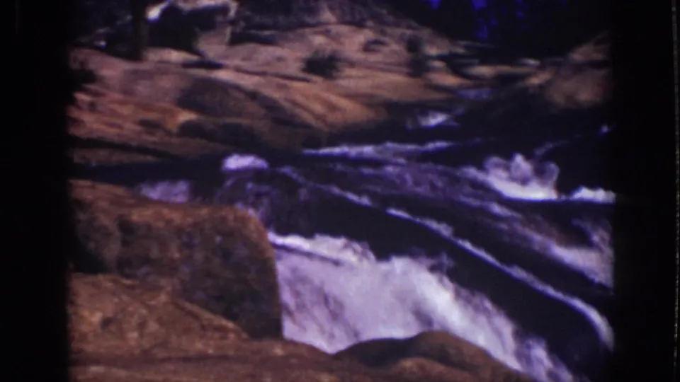 1939 Lake Tahoe home-movie frame of foaming waterfall water flowing between large rocks and forest shadows.