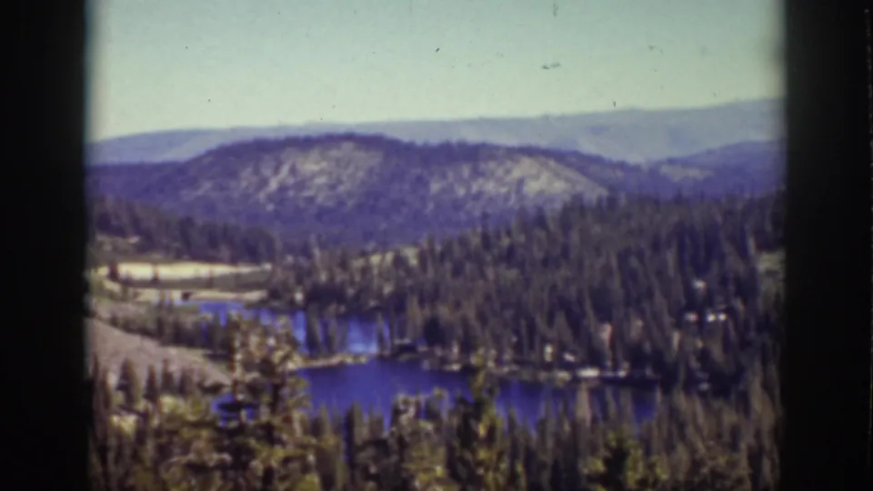 1939 Lake Tahoe panoramic view with blue water, dense conifers, rolling mountain slopes, and a pale sky.