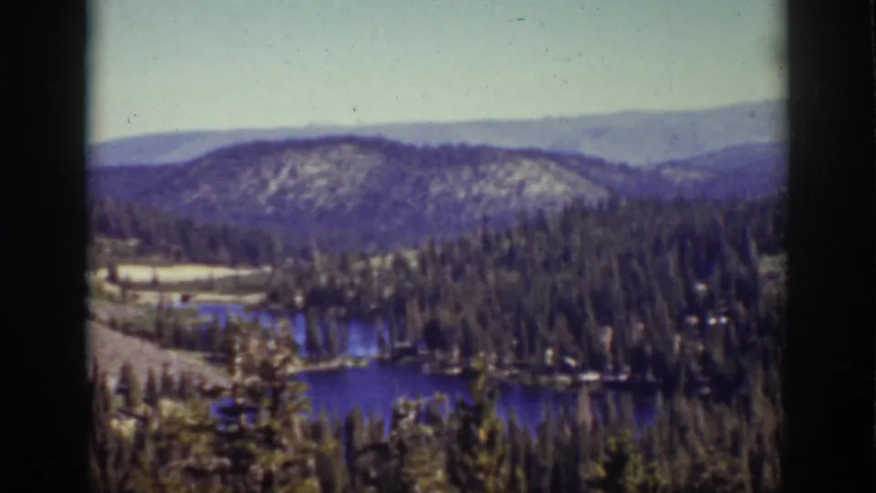 8mm Lake Tahoe landscape frame showing a blue lake below pine-covered slopes and layered hills in the distance.