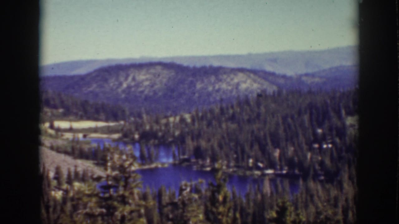 Blue lake among evergreen trees with mountain ridges in the distance.