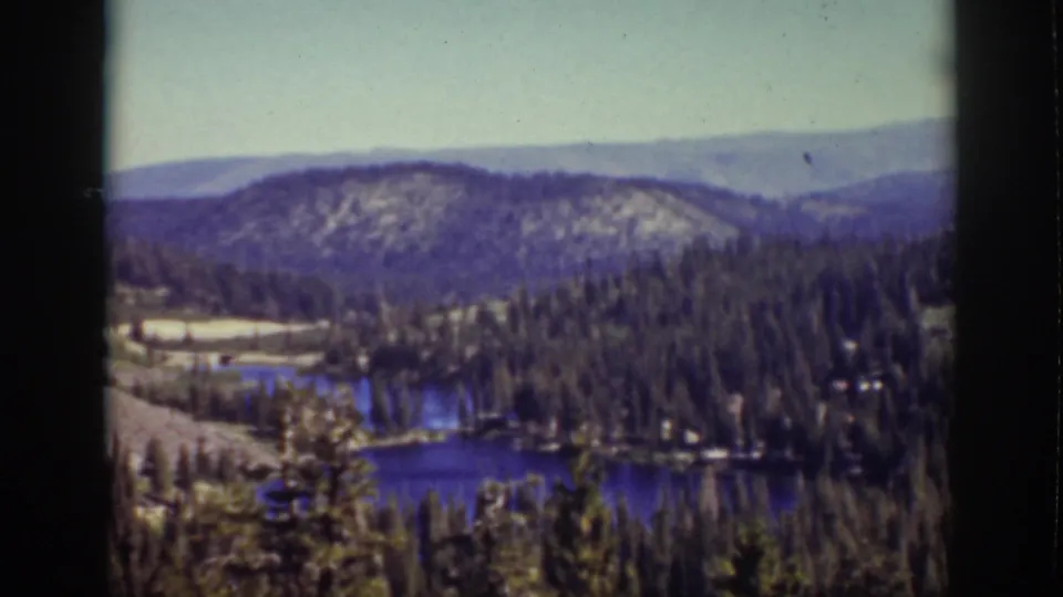 Soft-focus 1939 Lake Tahoe-area frame with blue lake water, evergreen forest, and a rounded hillside in the distance.