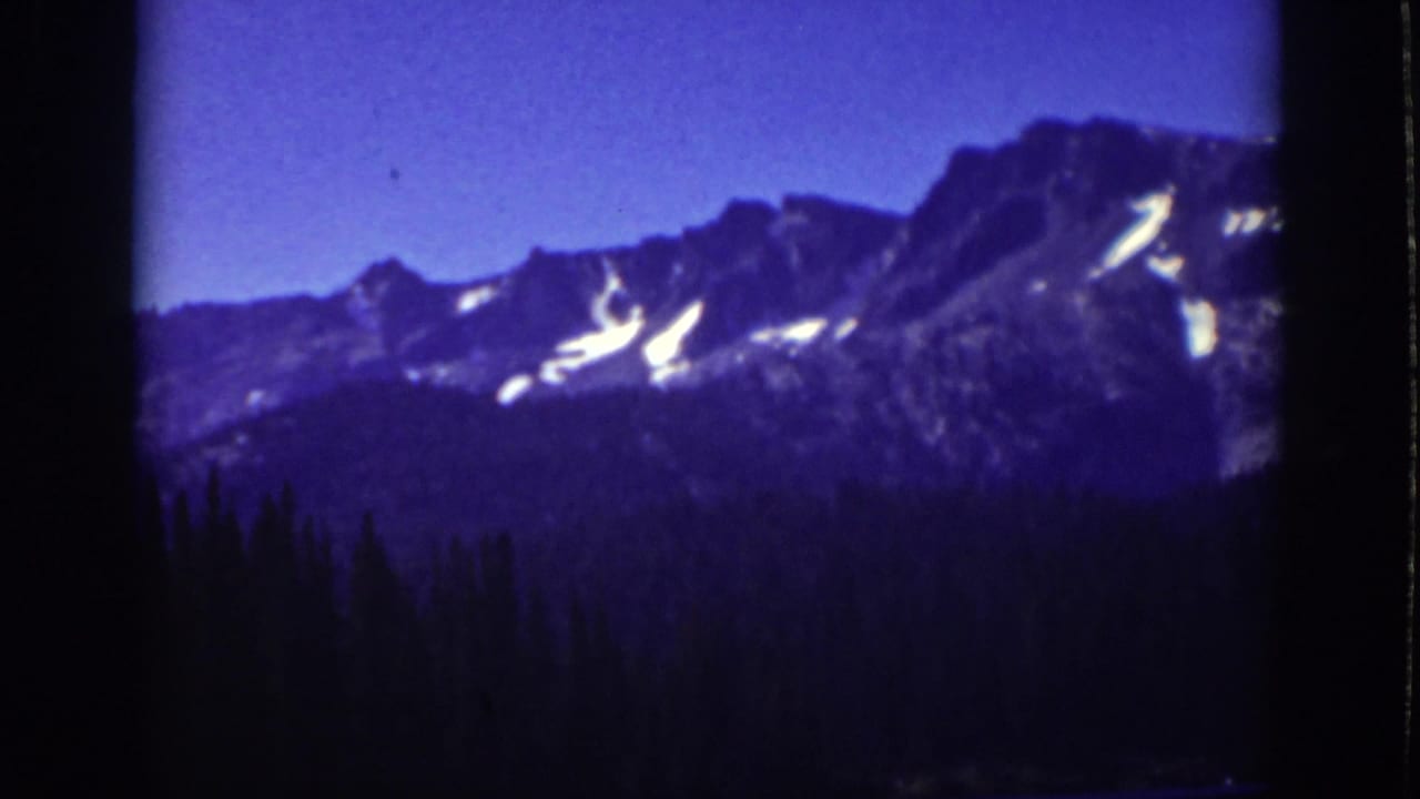 Snow-streaked mountain ridge above a dark evergreen forest under blue sky.