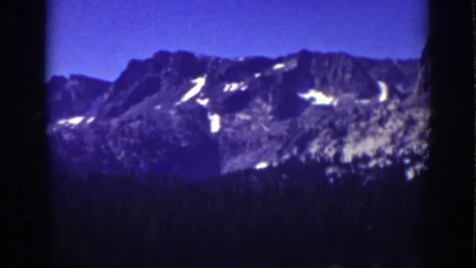 1939 Lake Tahoe 8mm frame showing a rocky Sierra Nevada ridge, small snow patches, blue sky, and dark trees below.