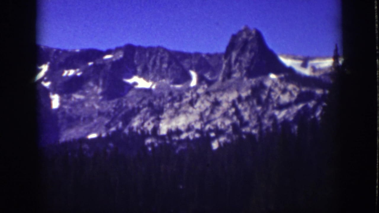 1939 Lake Tahoe 8mm frame showing distant rocky peaks with snow patches beyond a dark pine foreground and blue sky.