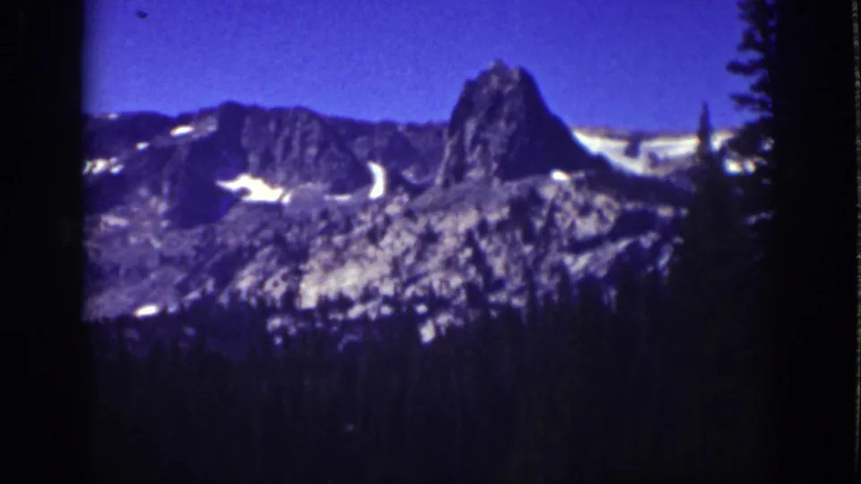 1939 Lake Tahoe 8mm frame showing a prominent rocky peak, snow-streaked ridge, blue sky, and shadowed pine forest below.