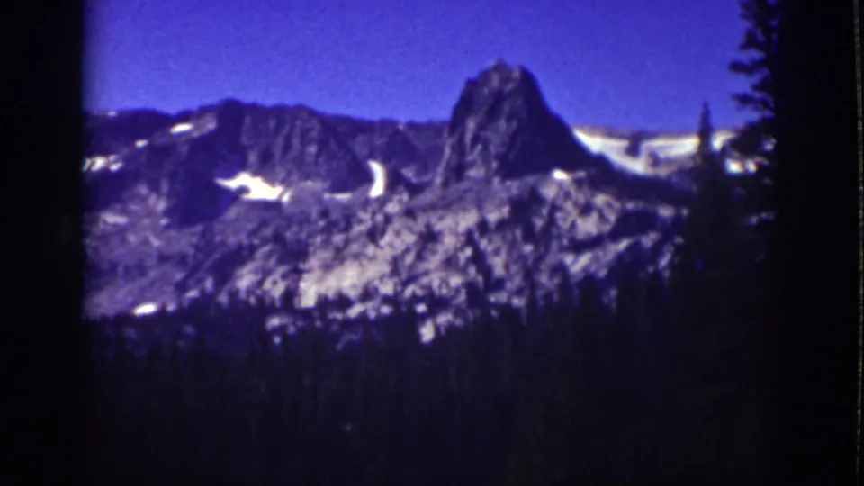 1939 Lake Tahoe scenic frame with soft-focus mountain peaks, scattered snow patches, dark pine edges, and saturated blue-purple color.
