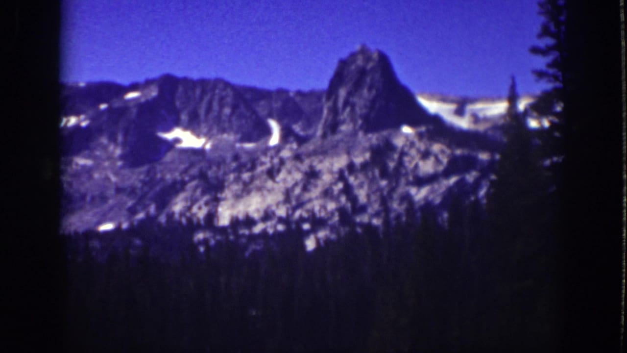 Snow-patched mountain peaks above a dark pine forest under blue sky.