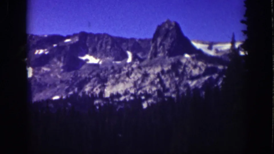 Final 1939 Lake Tahoe frame with a snow-patched rocky peak, saturated blue sky, and a dark evergreen lower edge.