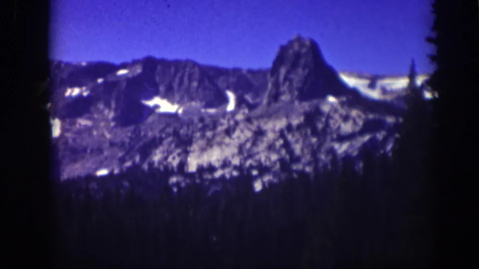 Vintage 8mm Lake Tahoe view with a blue-cast sky, snow-marked mountain slopes, and evergreen trees below.