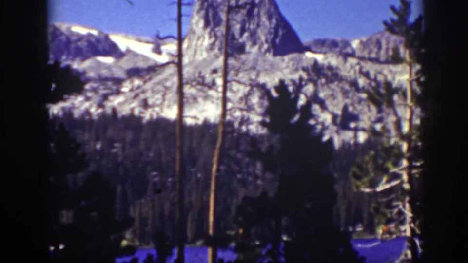 1939 Lake Tahoe-area home-movie frame with a rocky peak, snow patches, blue sky, and shadowed pine foreground.