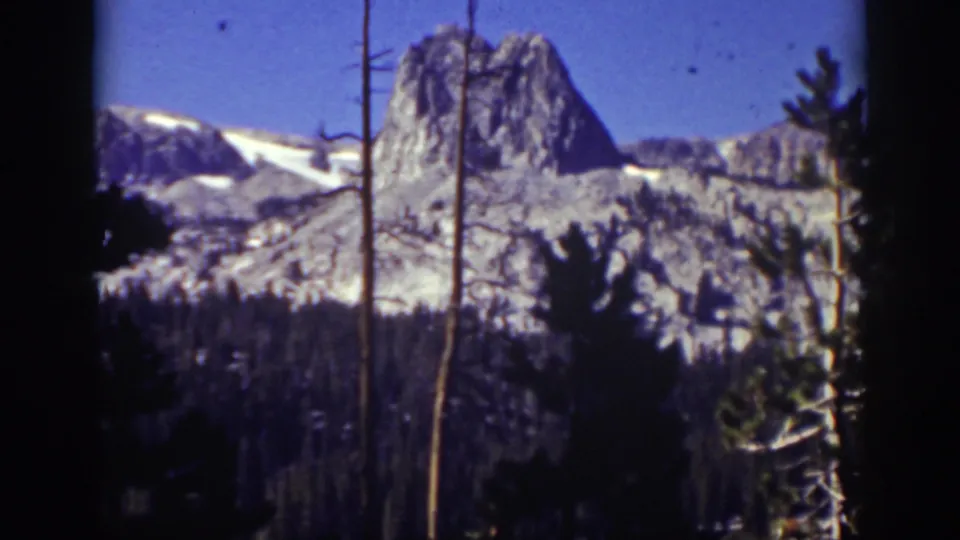 1939 Lake Tahoe-area 8mm frame showing a tall rocky peak above forested slopes, crossed by foreground tree trunks.